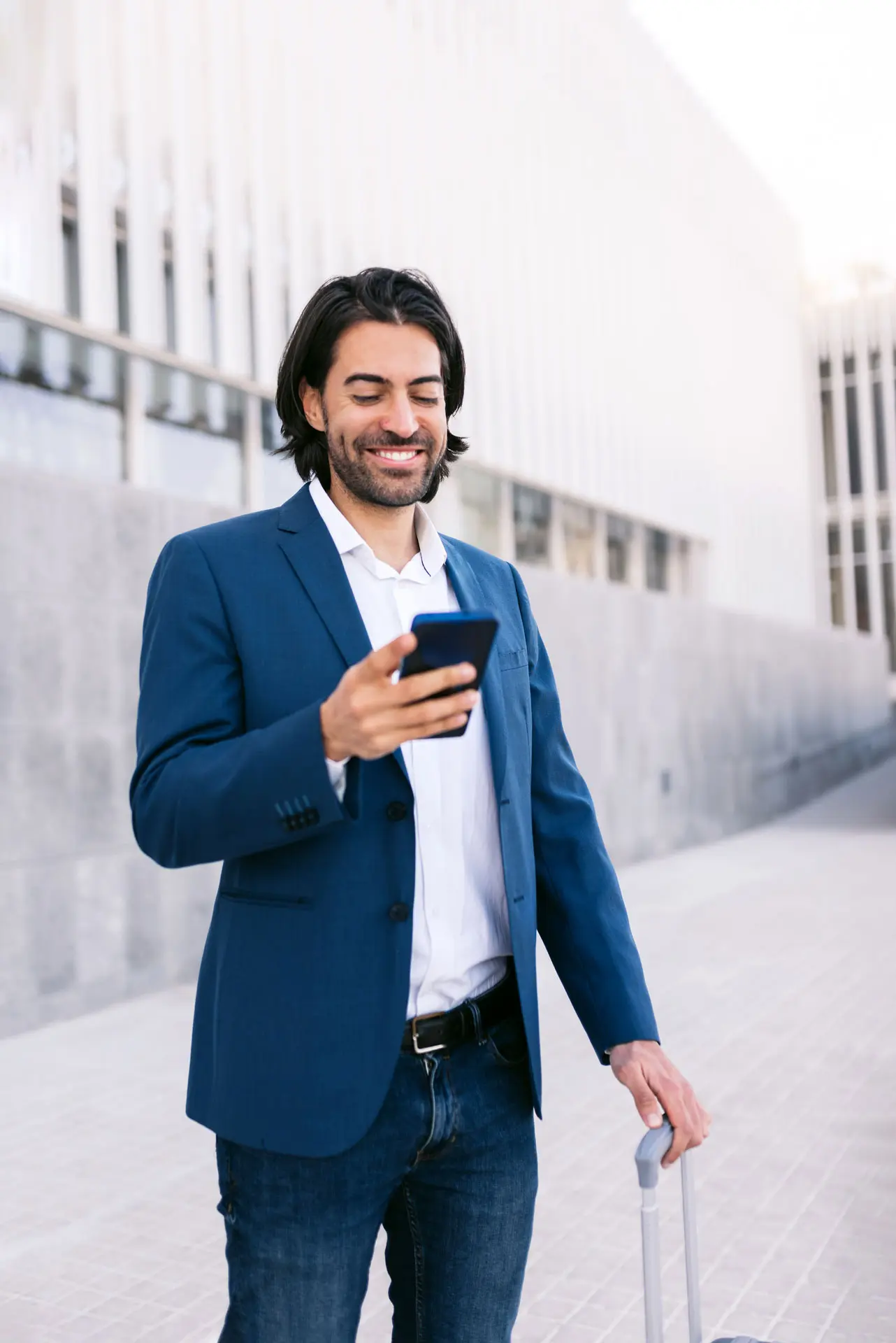 Geschäftsmann mit Rollkoffer lächelt beim Blick auf sein Handy, eventuell nach einer Wurzelbehandlung.